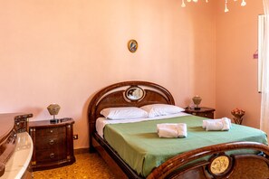 Ein ordentlich gemachtes Bett mit einem grünen Überwurf, zwei weißen Kissen und zwei Handtüchern. Ein Nachttisch aus Holz mit einer Lampe und einem kleinen dekorativen Gegenstand steht daneben. Das Zimmer verfügt über ein Holz-Kopfteil und eine Holzkommode mit Schubladen.