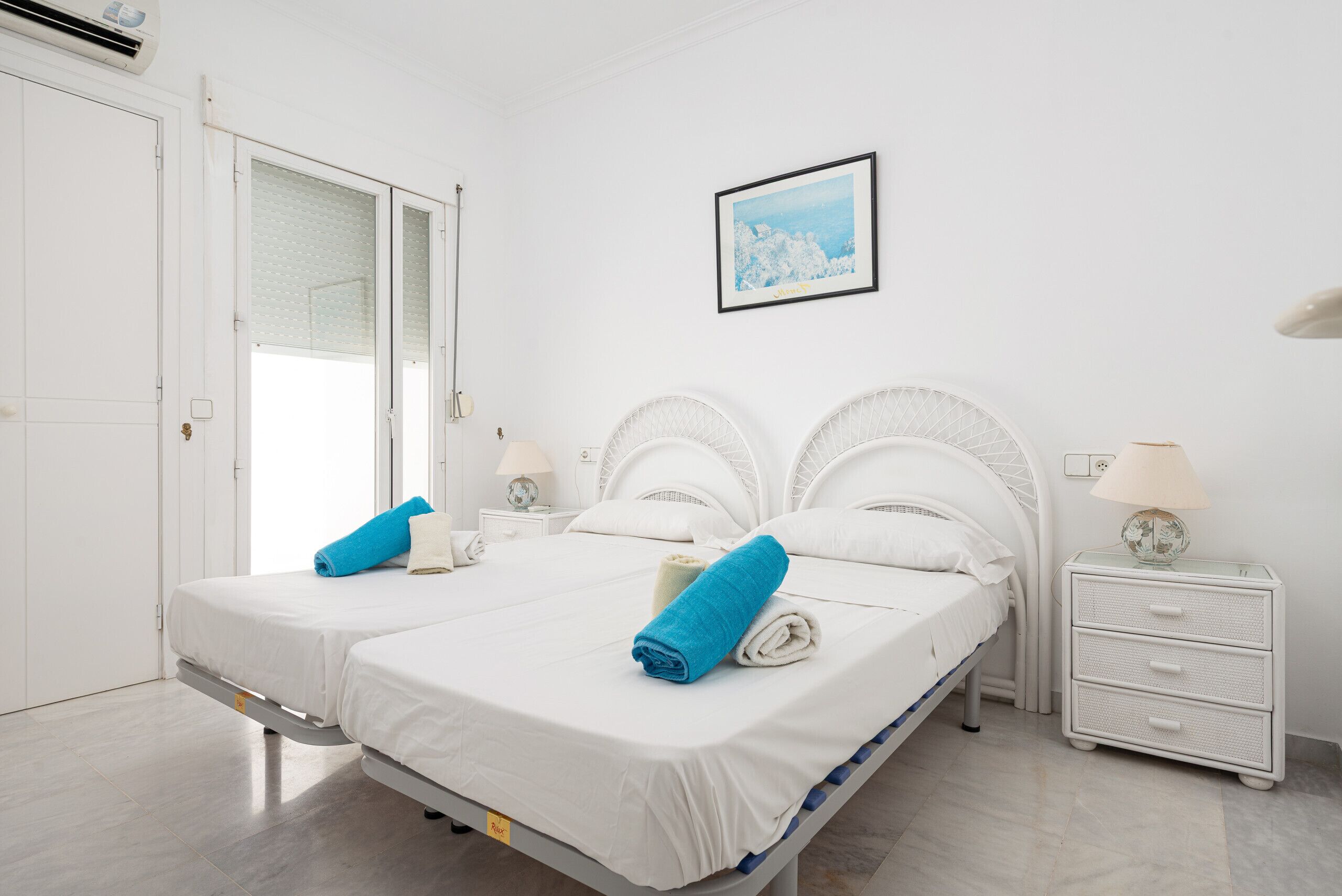 Una cama doble con ropa de cama blanca y toallas azules, una mesita de noche con una lámpara y un cuadro en la pared.