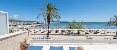 Un balcon avec des fauteuils de jardin, un palmier et une vue sur une plage où l’on aperçoit des bateaux et des parasols.