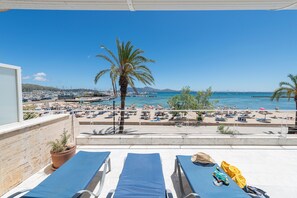 A balcony with lounge chairs, a palm tree, and a view of a beach with boats and umbrellas. - Port de Pollen a Topacio 1 (Pollença)