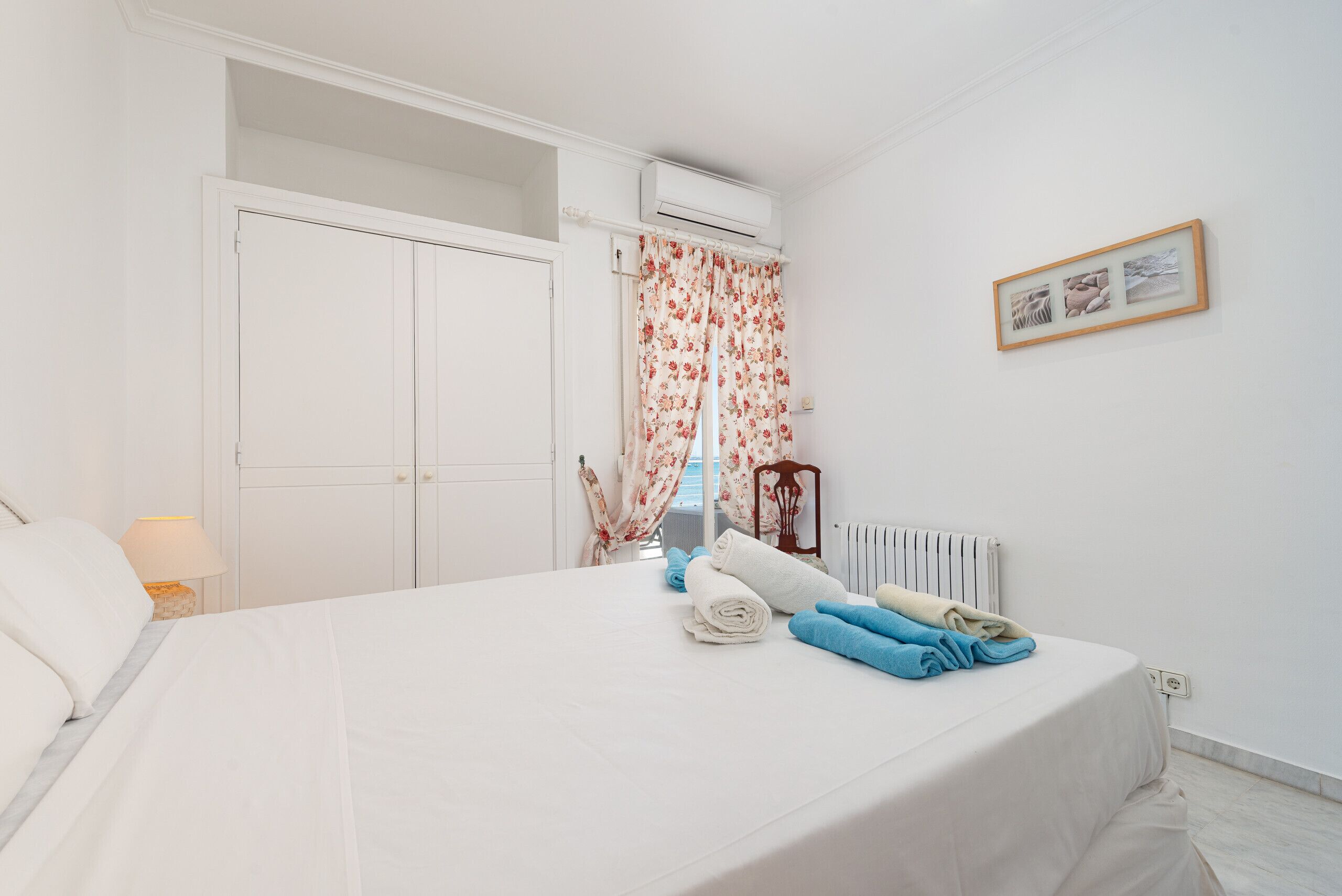 A bedroom with a bed, a radiator, a window with floral curtains, a framed picture on the wall, and a white door.