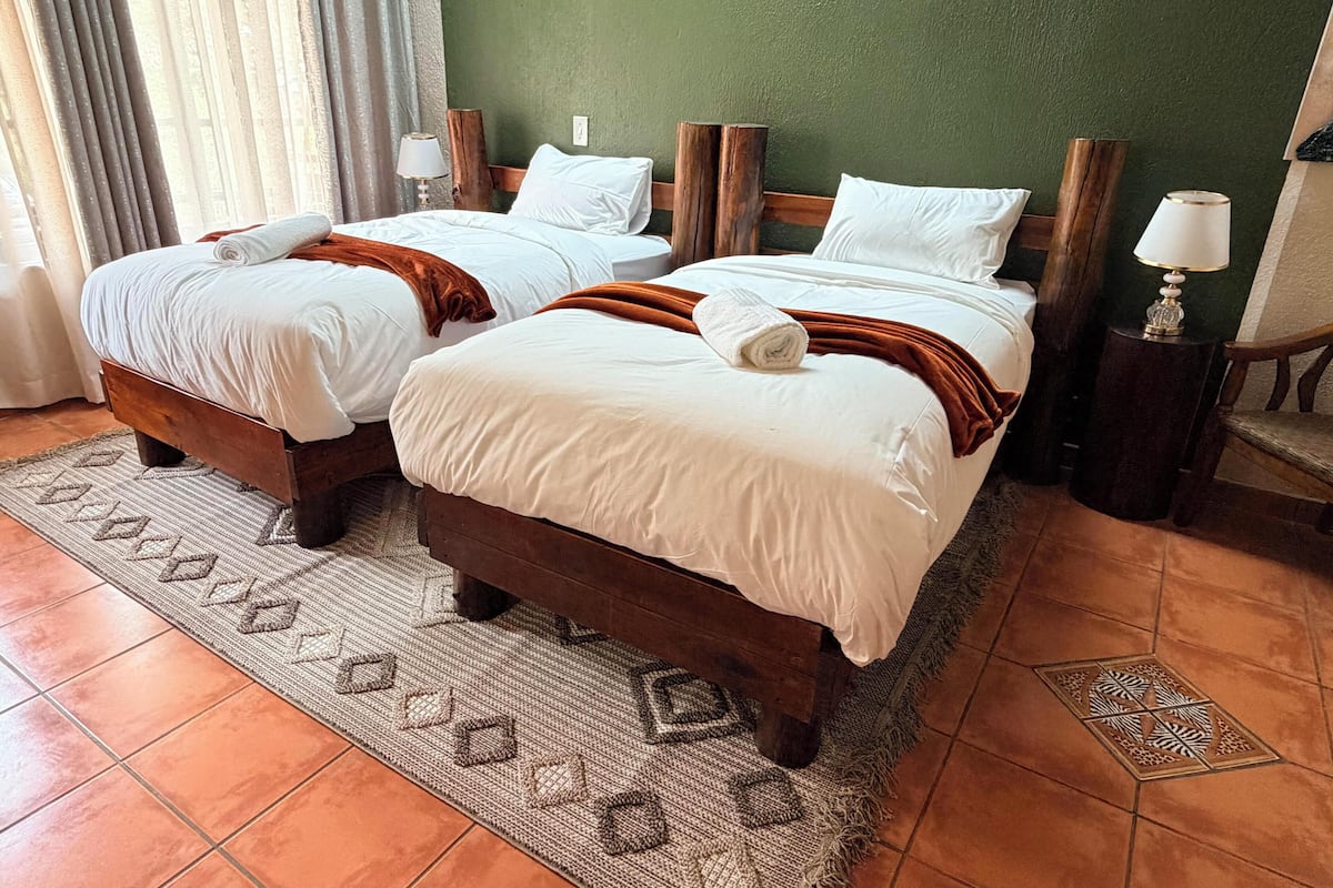 Dos camas individuales con cabeceros de madera, ropa de cama blanca y una alfombra tejida sobre suelo de baldosas.