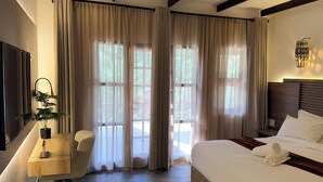 A hotel room with a bed, a desk, a chair, a potted plant, and large windows with curtains. - Mvubu Falls Lodge (Lobamba)