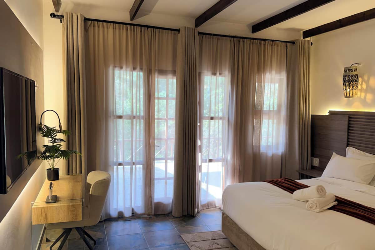 Habitación de hotel con cama, escritorio, una silla, planta en maceta y ventanas grandes con cortinas.