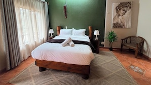 A hotel room with a large bed, a wooden headboard, a green accent wall, a painting, a chair, and a window with curtains. - Mvubu Falls Lodge (Lobamba)