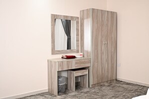 Een slaapkamer met een houten garderobe, een spiegel, een klein kaptafeltje met een rode tas en een prullenbak.