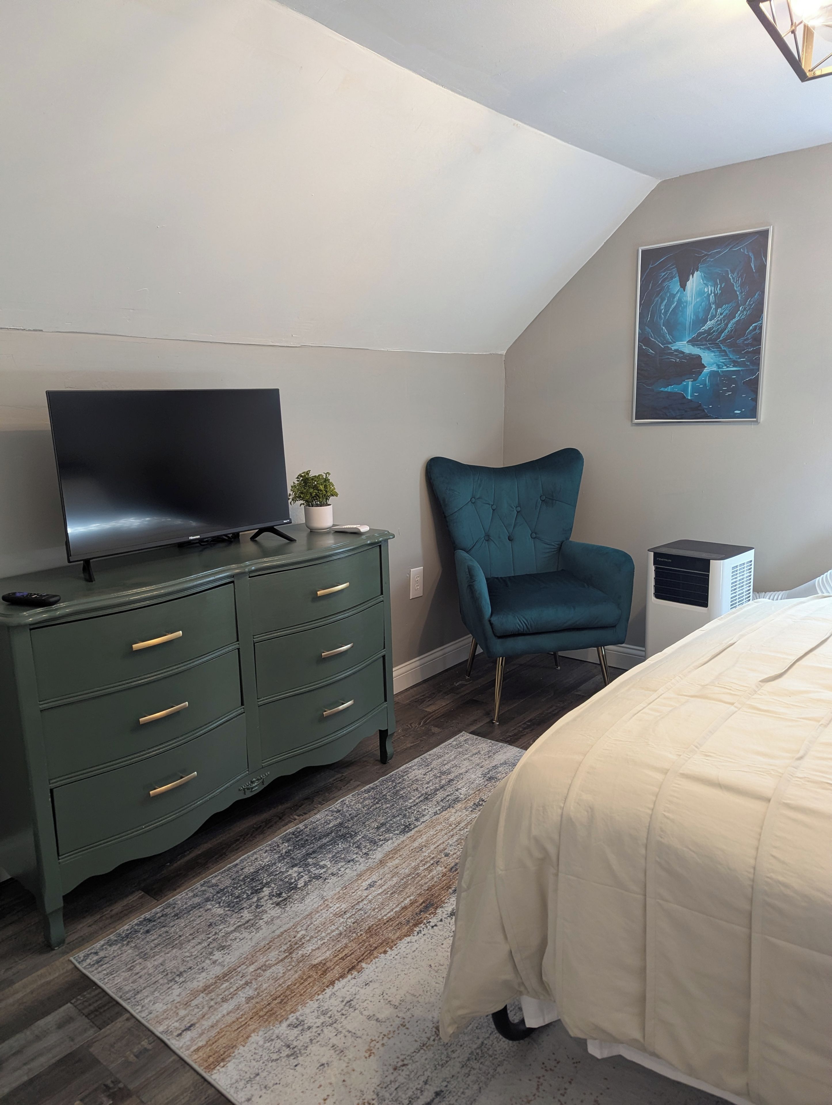 Ein Schlafzimmer mit einem Bett, einem Fernseher auf einer Kommode, einem Sessel und einem Bild an der Wand.
