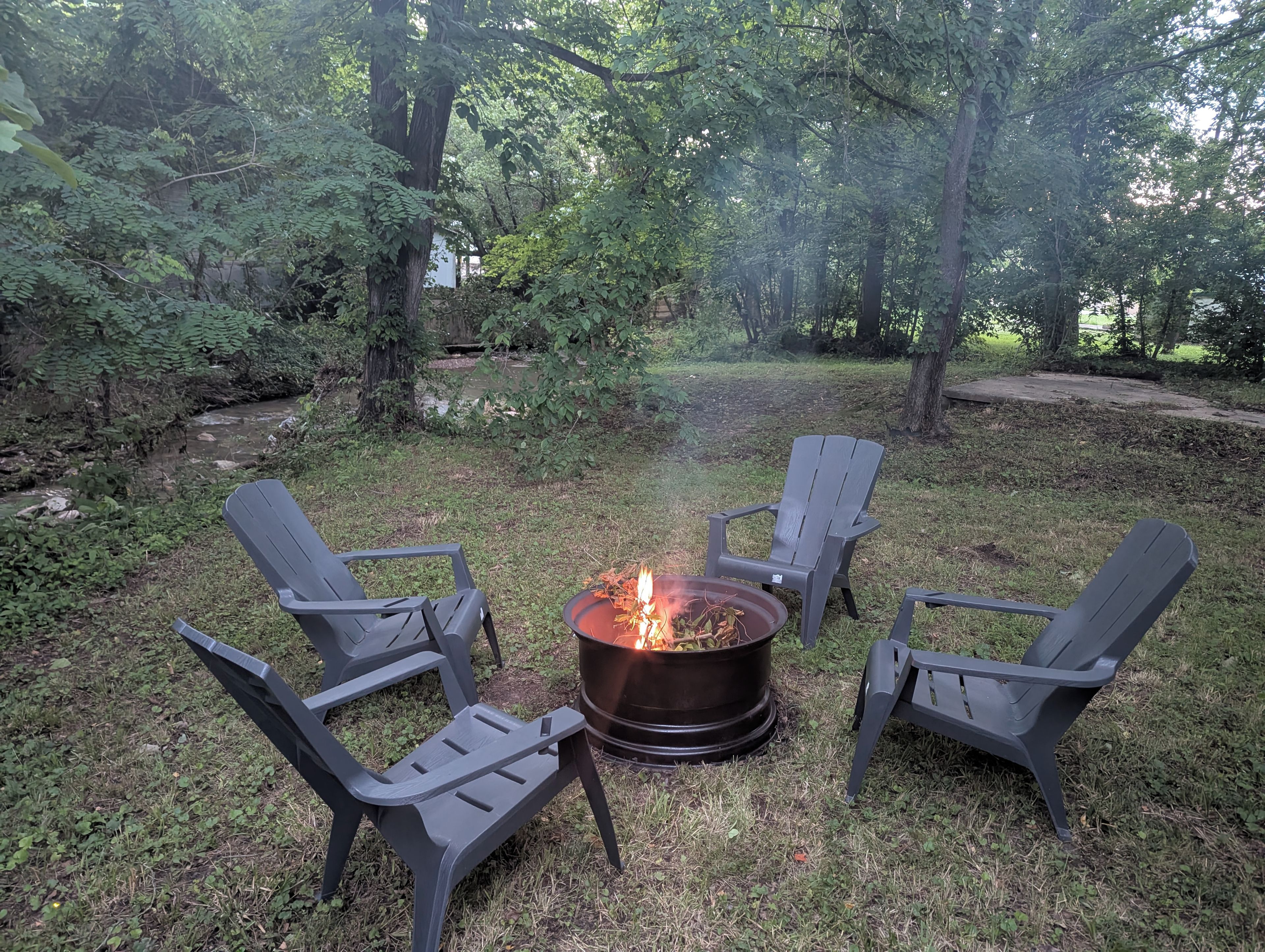 Un foyer extérieur entouré de quatre chaises Adirondack, le tout installé dans un espace boisé.