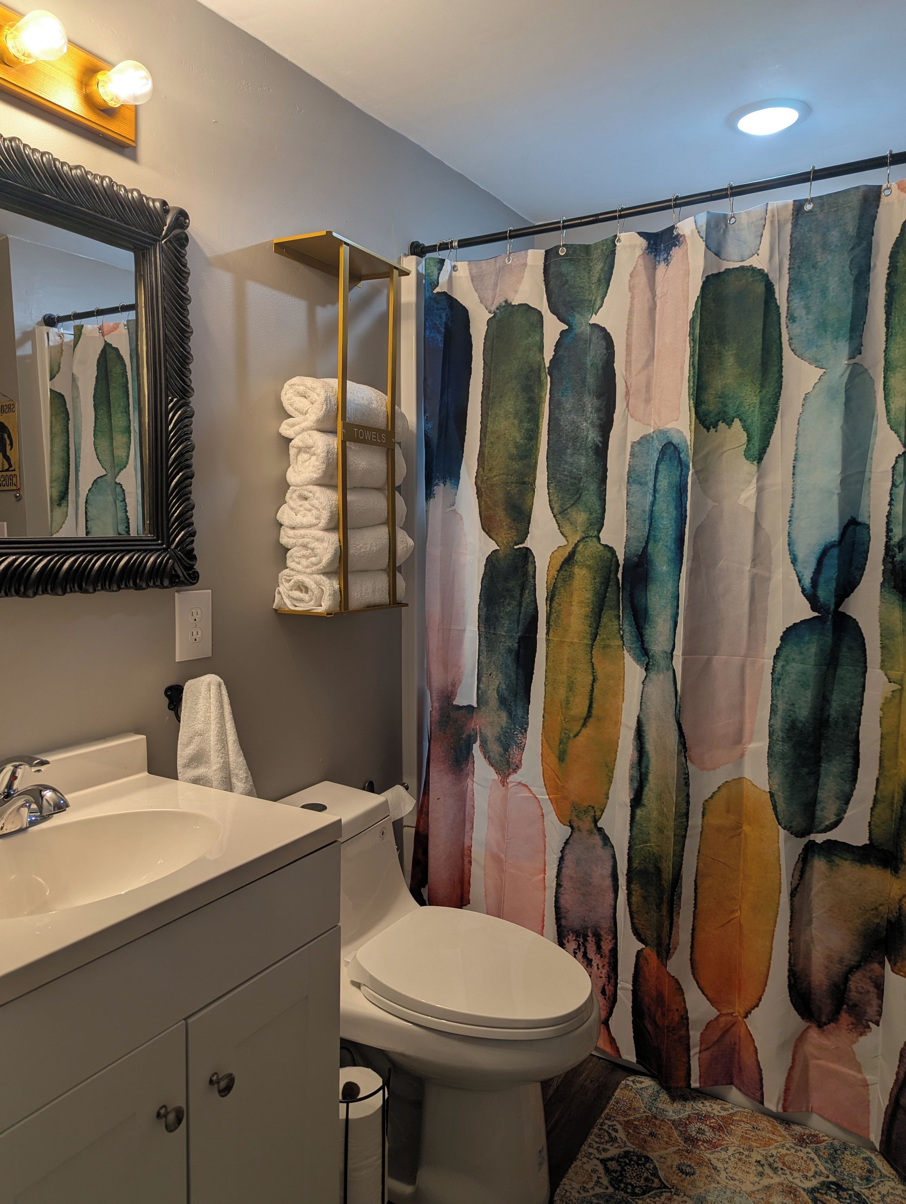 Un bagno con lavandino, water e box doccia con decoro acquerello. Si vede uno scaffale portasciugamani con asciugamani piegati e uno specchio con luci sopra.