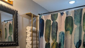 A bathroom with a white sink, toilet, and a shower curtain with a watercolor design. There is a towel rack with folded towels and a mirror with lights above it. - Cave Country Creekside Getaway (Marengo)