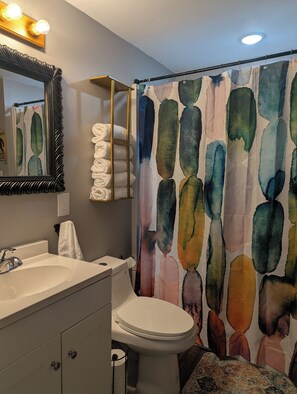 A bathroom with a white sink, toilet, and a shower curtain with a watercolor design. There is a towel rack with folded towels and a mirror with lights above it. - Cave Country Creekside Getaway (Marengo)