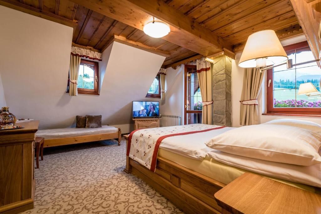 Un dormitorio acogedor con techo de madera, una cama con ropa de cama blanca, una mesita de noche, un televisor y una ventana con cortinas.