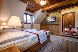 A bedroom with a bed, a TV mounted on the wall, wooden ceiling beams, and a small table with glasses and a bottle.
