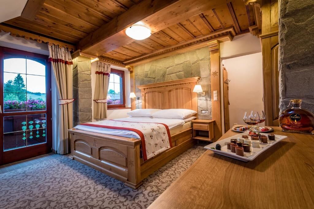 A cozy bedroom with a wooden bed, a stone wall, a window with curtains, and a tray of chocolates.
