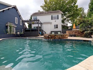 Una piscina residencial con un muro de acento de piedra, un pérgola y una casa de dos pisos al fondo.