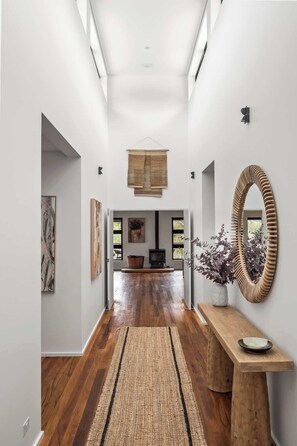 A hallway with a wooden floor, a woven rug, a wooden bench with a plate, a wall-mounted mirror, and a fireplace. - The Orchard—Refined Living & Natural Serenity (Margaret River)