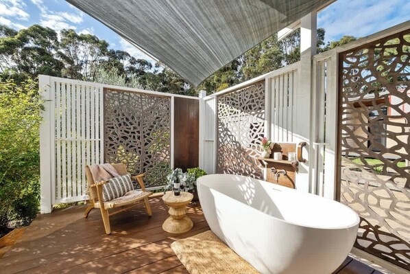 Un patio exterior con una bañera blanca, una silla de madera y una mesa con una botella.