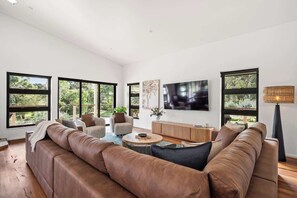 A modern living room with a brown leather sofa, a flat-screen TV, and large windows offering a view of greenery.