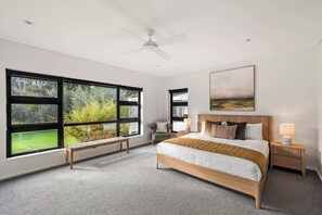A bedroom with a large bed, a bench, a nightstand, a ceiling fan, and a window with a view of greenery.