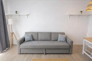 A grey sofa with patterned cushions, a wooden floor, a white lamp, and two white shelves with small plants.