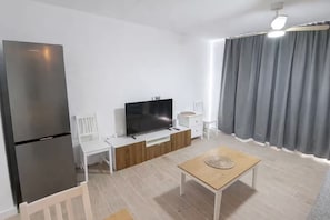 A modern living room with a flat-screen TV, a wooden coffee table, a white sofa, and a grey curtain. - Oasis Duna-Pool Side View Apartment-Corralejo (Corralejo)