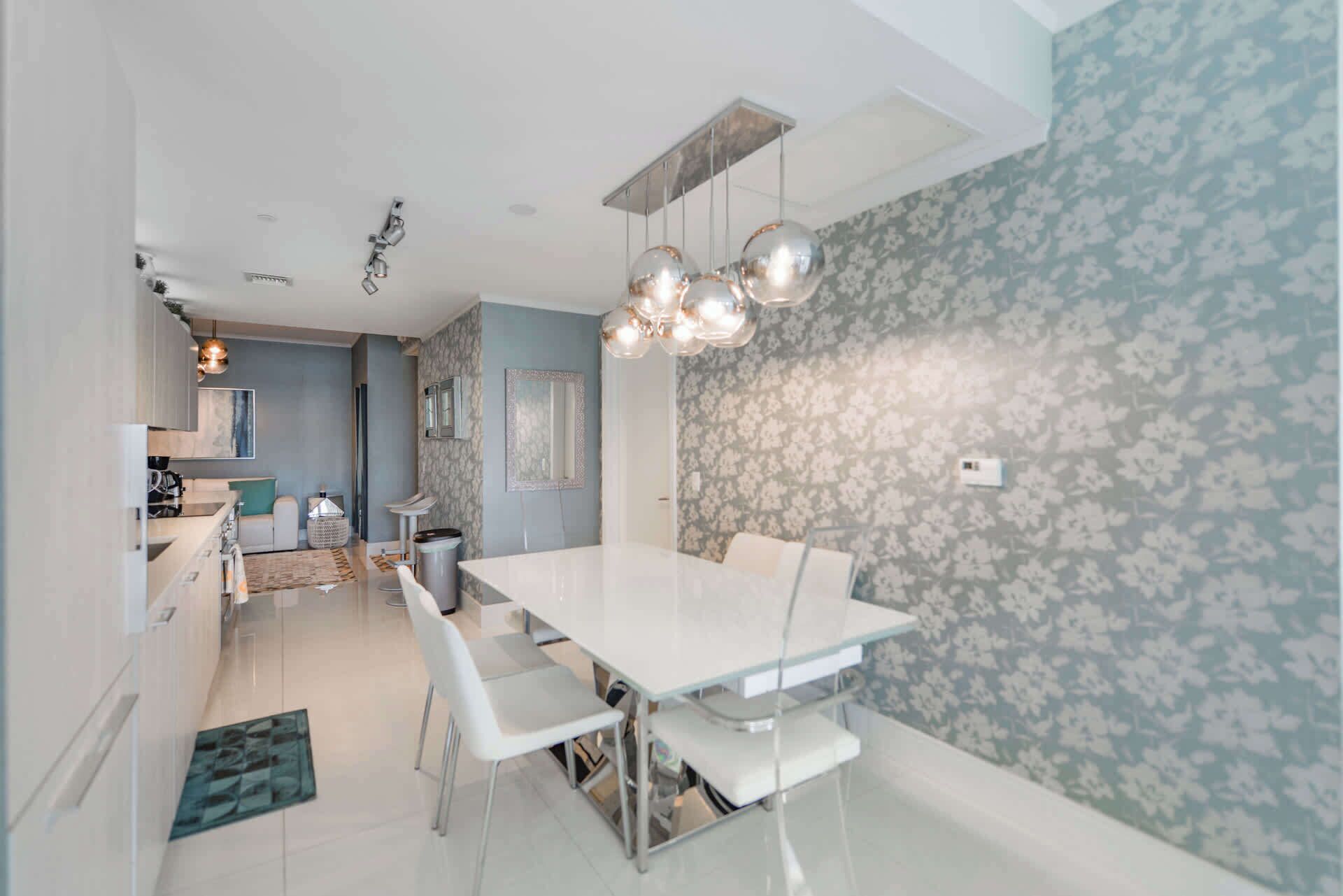 A modern dining area with a white table and chairs, a patterned wall, and a kitchen in the background.