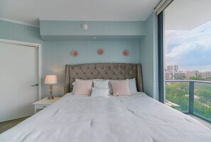 Une chambre à coucher avec un grand lit, une table de chevet, une lampe et une vue sur les bâtiments par la fenêtre.