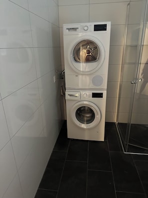 A stacked washer and dryer unit in a bathroom with white tiled walls and a black tiled floor.