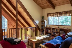 A cozy loft-style room with a wooden ceiling, a balcony with a railing, a bookshelf, a sofa, and a window with a view of trees. - Two Incredible Cabins for a Memorable Family Getaway in Dorrington, California (Dorrington)