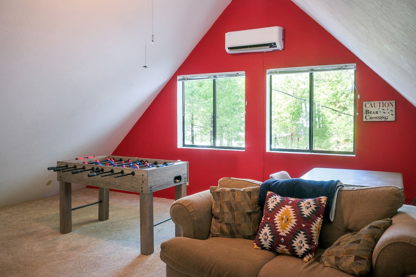 A room with a foosball table, a couch with patterned cushions, and two windows with a view of greenery.