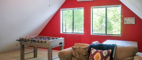 A room with a foosball table, a couch with patterned cushions, and two windows with a view of greenery.