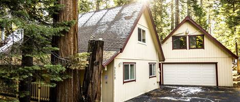 A two-story house with a garage, surrounded by tall trees and a wooden deck.