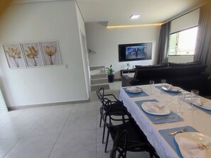 A dining area with a table set for four, a flat-screen TV mounted on the wall, and a staircase leading to an upper level. - New, cozy 3-bedroom townhouse near the sea and the center of Matinhos. (Matinhos)