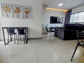 A modern living room with a black sofa, a TV mounted on the wall, a dining area with two chairs, and a staircase leading to an upper level. - New, cozy 3-bedroom townhouse near the sea and the center of Matinhos. (Matinhos)