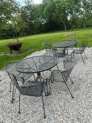 Ein Outdoor-Esstisch-Set aus Metall mit rundem Tisch und Stühlen, umgeben von Kies, mit einer Rasenfläche und Bäumen im Hintergrund.