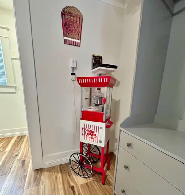 A corner with a red and white popcorn cart, a white wall, a window with blinds, and a wooden floor.