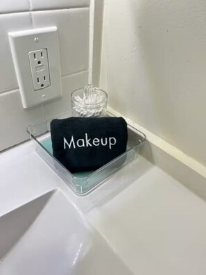 A bathroom with a sink, a towel rack with a black towel labeled 'Makeup,' and a glass container on the counter.