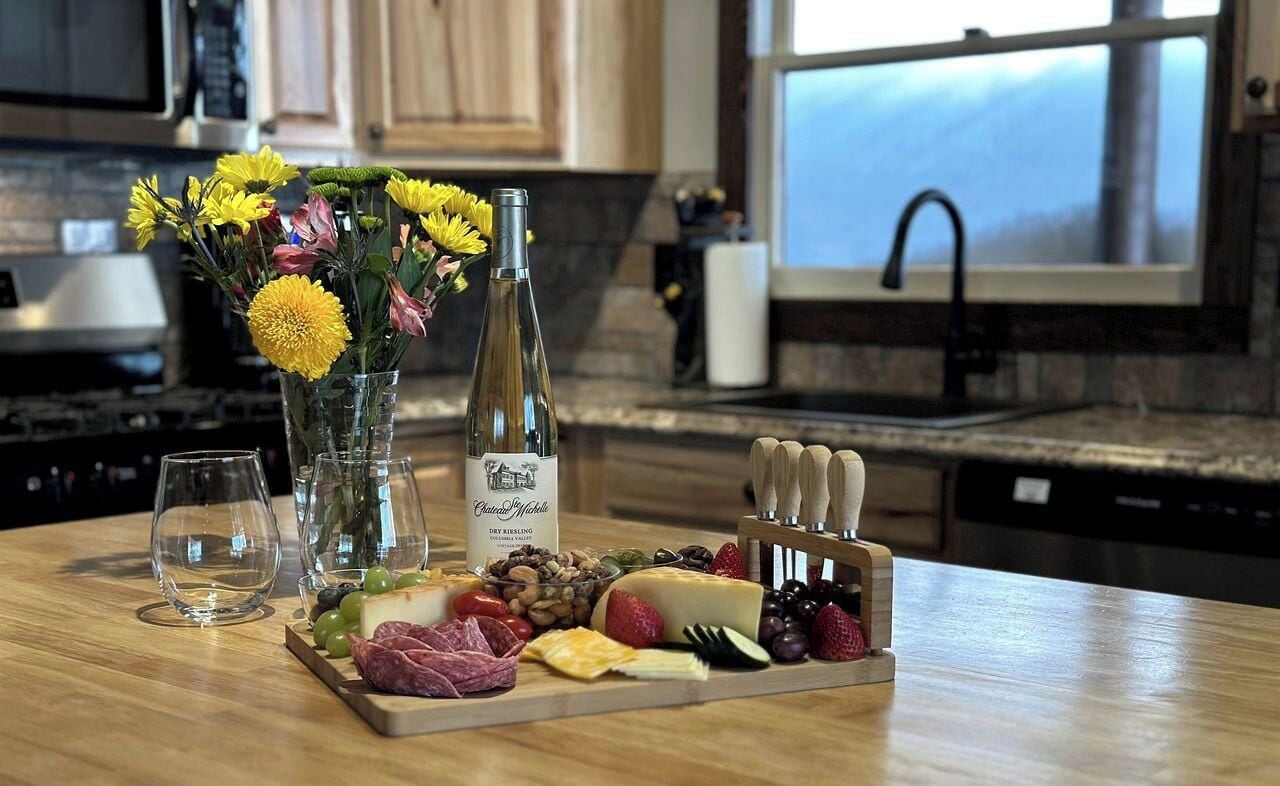 A kitchen counter with a wine bottle, glasses, and a cheese board with various cheeses and fruits.