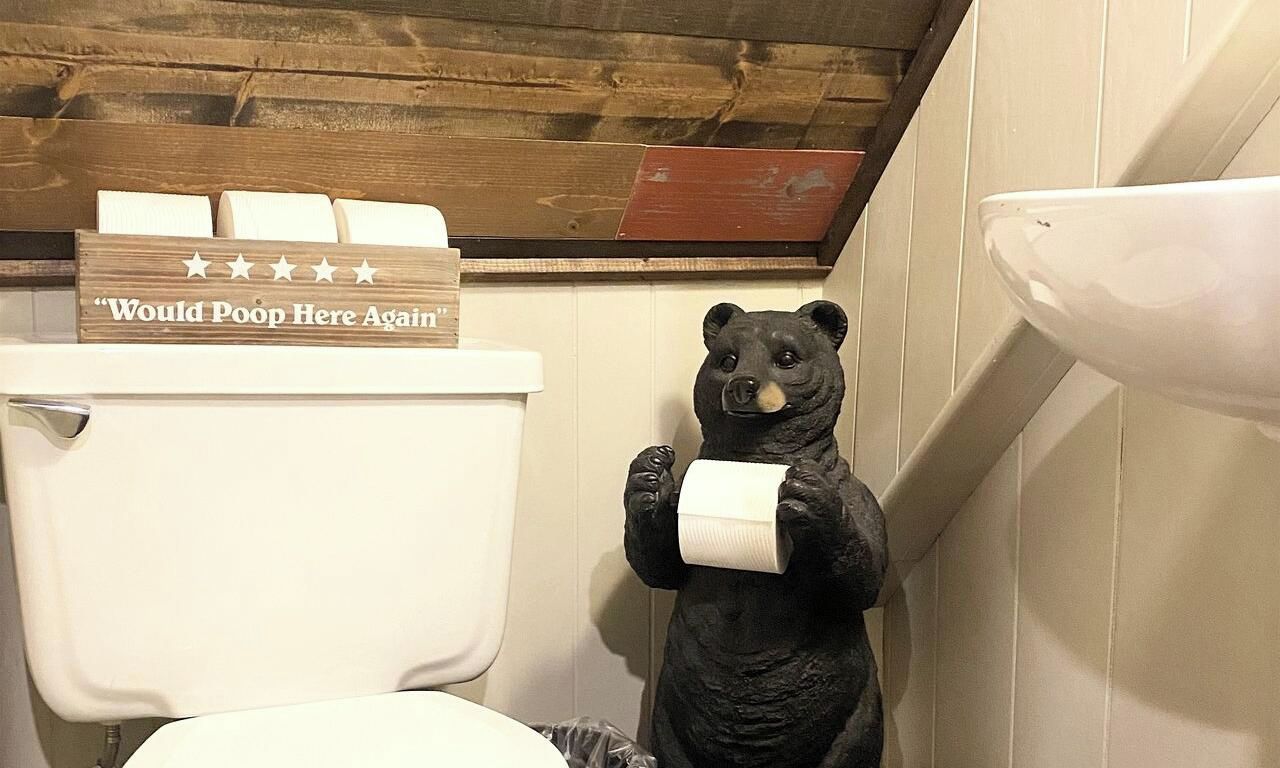 A bathroom with a toilet, a bear statue holding a roll of toilet paper, and a sign above the toilet that reads 'Would Poop Here Again'.