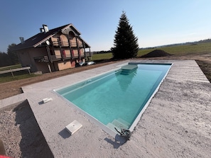 A rectangular swimming pool with a concrete deck, a house with a sloped roof, and a large evergreen tree in the background.
