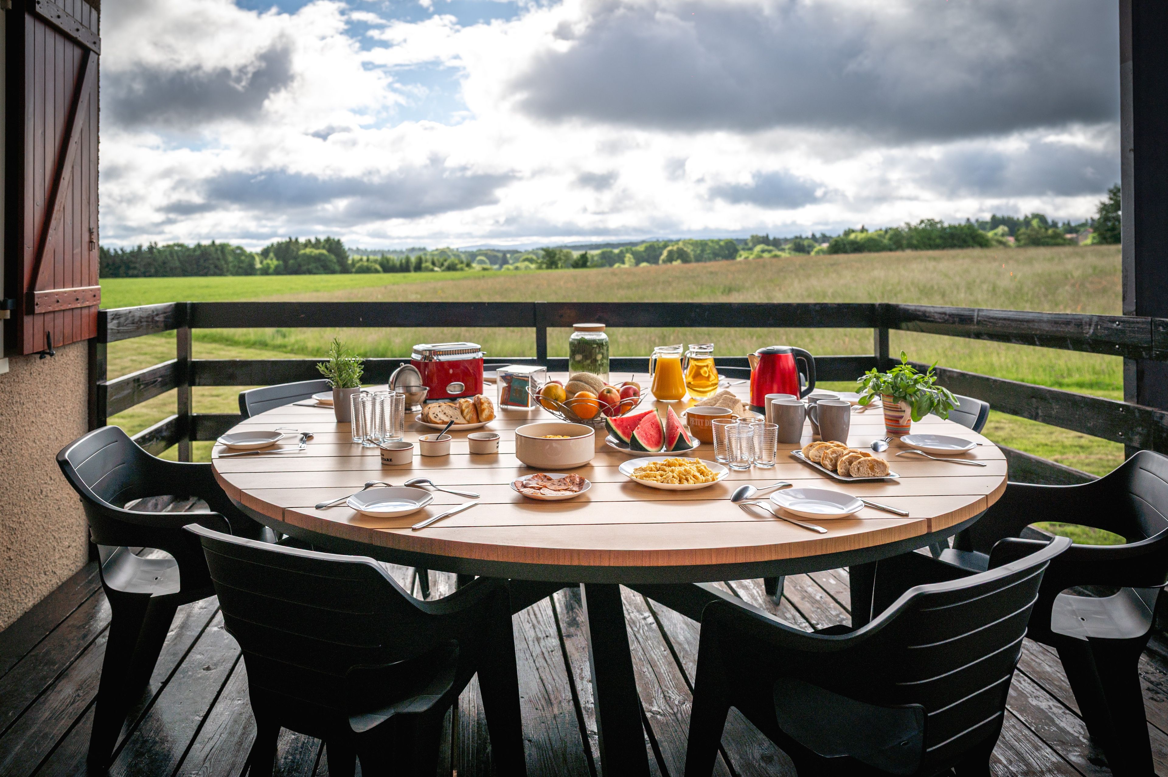 Une table ronde en bois, dressée pour le petit-déjeuner avec des assiettes, des tasses et une variété d’aliments, est placée sur un balcon donnant sur un champ verdoyant.