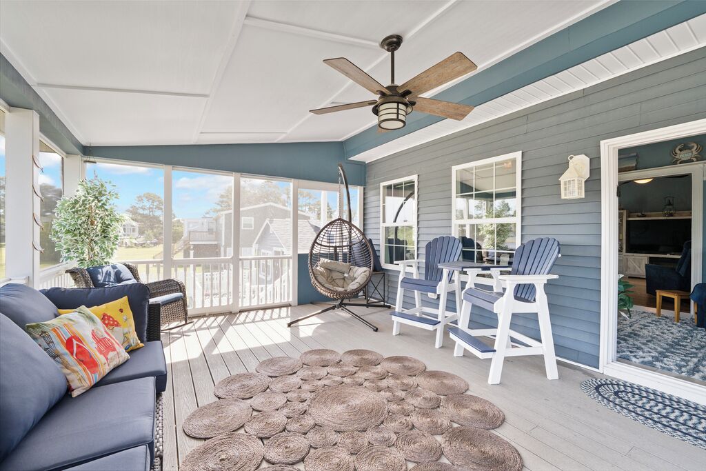 Une véranda couverte avec un ventilateur de plafond, un canapé bleu agrémenté de coussins colorés, un tapis tissé et des fauteuils Adirondack blancs.