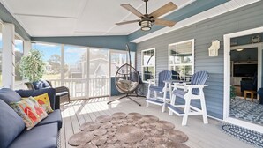 A covered porch with a ceiling fan, a blue sofa with colorful cushions, a woven rug, and white Adirondack chairs.