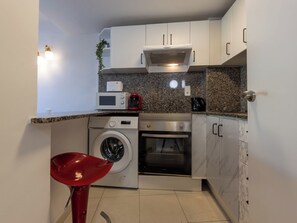 A compact kitchen with a microwave, oven, and washing machine, a red chair, and white cabinets. - Apartment Royal by Interhome (Salou)
