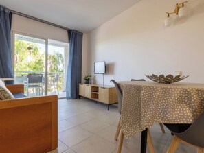 Ein Zimmer mit Esstisch, Stühlen, Fernseher, Schrank und einem Balkon mit Blick ins Grüne.