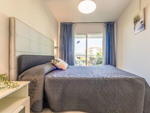 A bedroom with a bed, a bedside table, a headboard, a window with curtains, and a view of buildings outside. - Apartment Royal by Interhome (Salou)