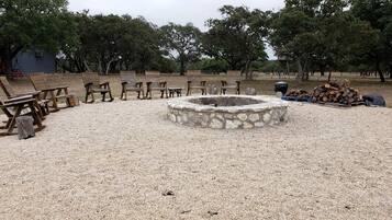 A gravel area with wooden chairs and benches arranged around a stone fire pit.