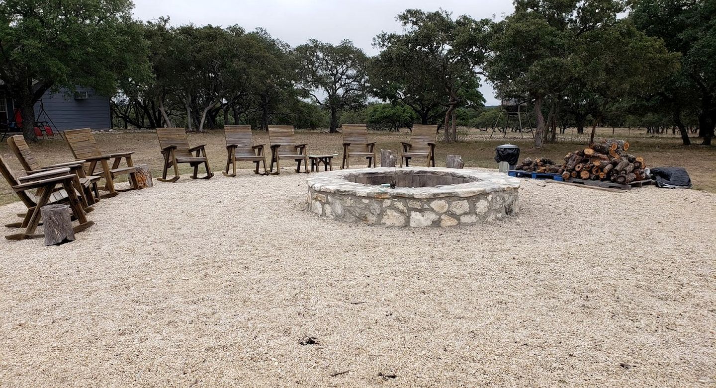 A gravel area with wooden chairs and benches arranged around a stone fire pit.