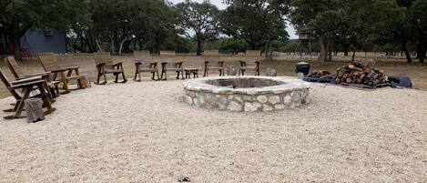 A gravel area with wooden chairs and benches arranged around a stone fire pit.