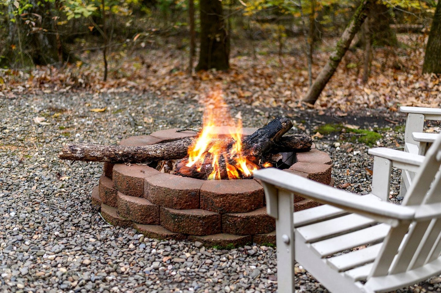 A circular brick fire pit with a log fire, surrounded by gravel, with a white chair in the foreground.